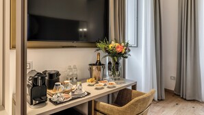 Une chambre d’hôtel moderne dotée d’une grande télévision à écran plat, d’un minibar, d’une table à manger avec des produits pour le petit-déjeuner, d’un fauteuil en osier et d’un vase rempli de fleurs.