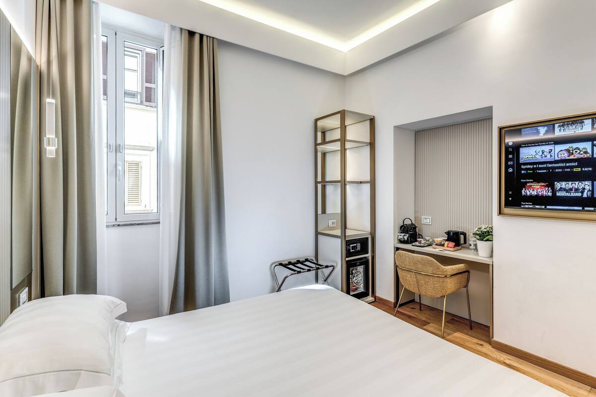 Una habitación de hotel con cama, escritorio con silla, televisor montado en la pared y ventana con cortinas.
