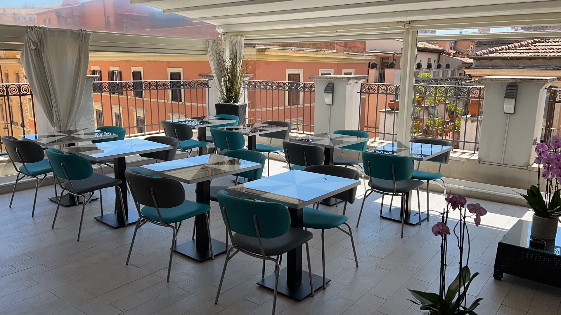 An outdoor cafe with tables and chairs, a potted plant, and a view of buildings.