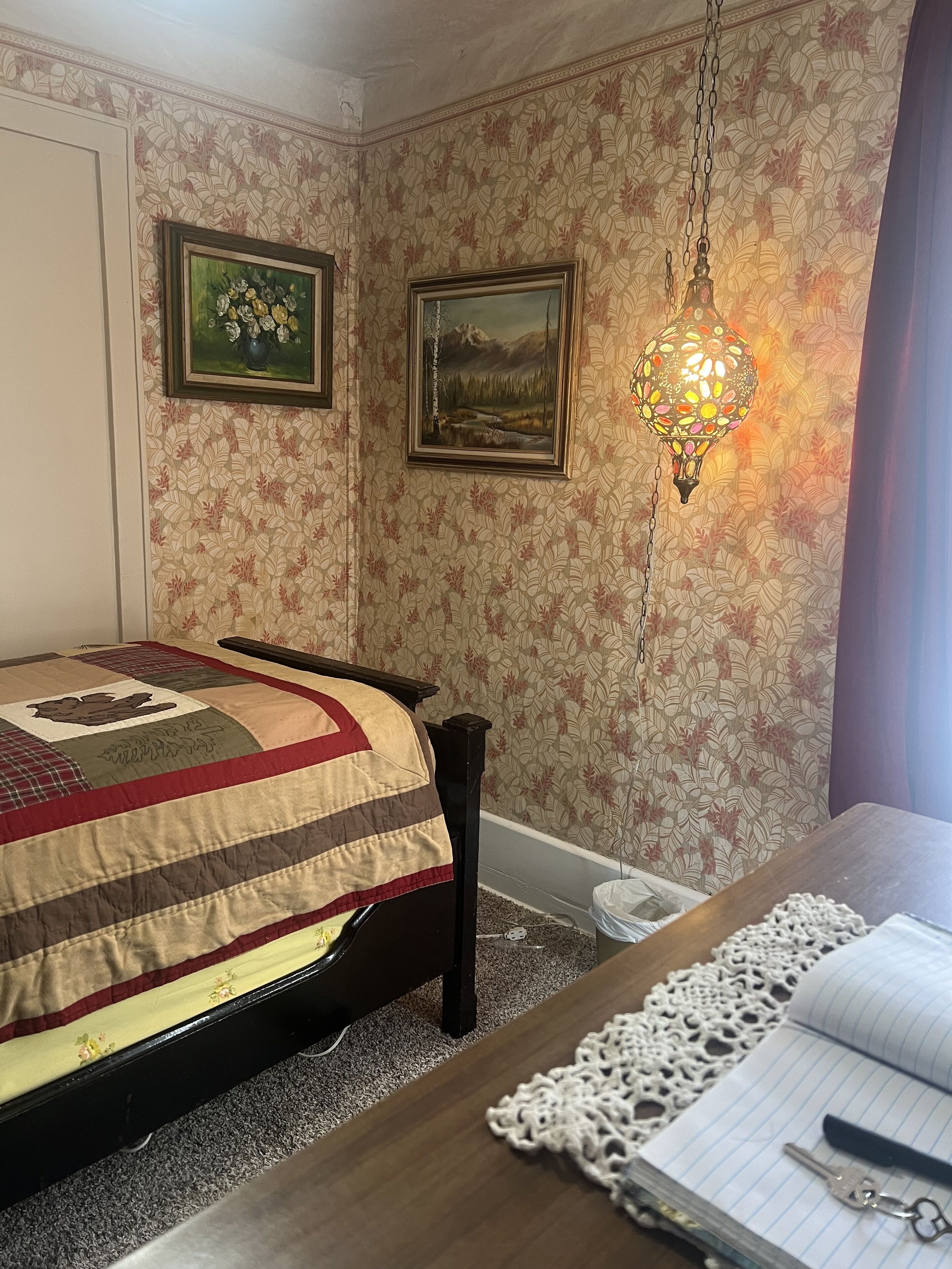 Une chambre à coucher avec un lit, une table de chevet en bois, un bureau avec un carnet, et deux tableaux encadrés accrochés au mur.