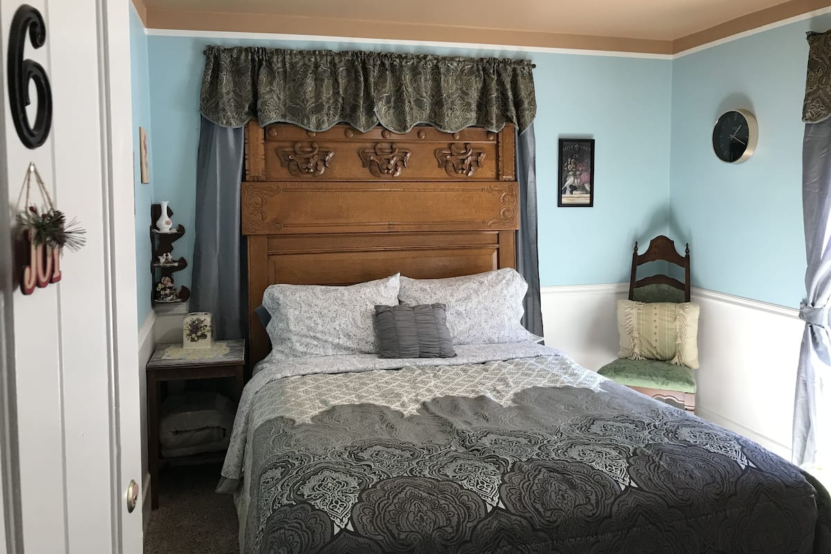 A bedroom with a wooden headboard, a patterned bedspread, a nightstand with a vase, and a clock on the wall.