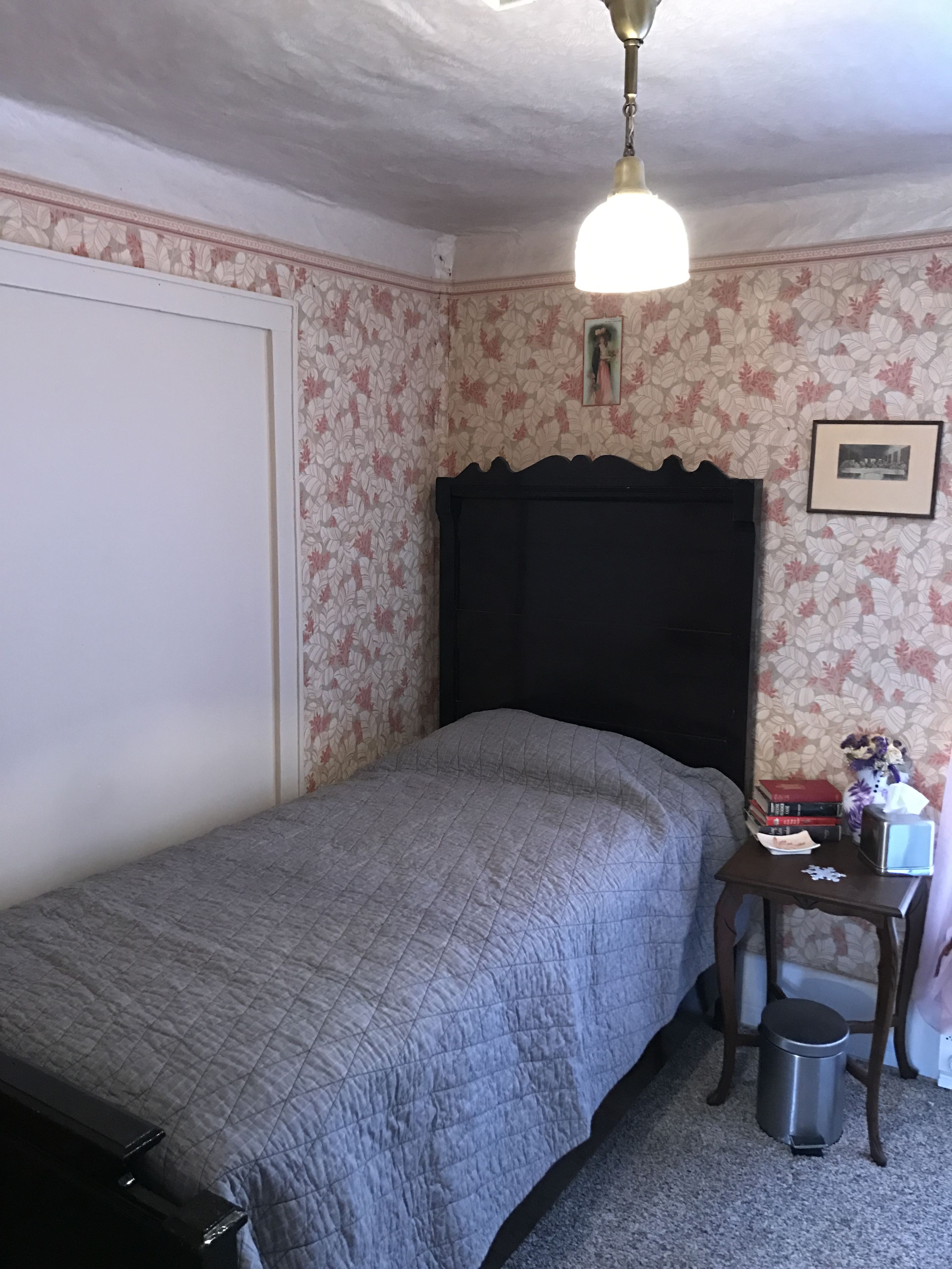 A bedroom with a single bed, a nightstand, a trash can, and a wall with floral wallpaper.