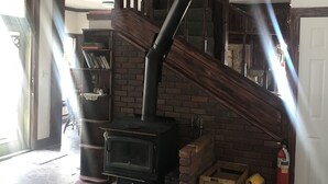 A wood-burning stove in a living room with a staircase and a fireplace. - Historic Windsor Hotel (Silver Plume)