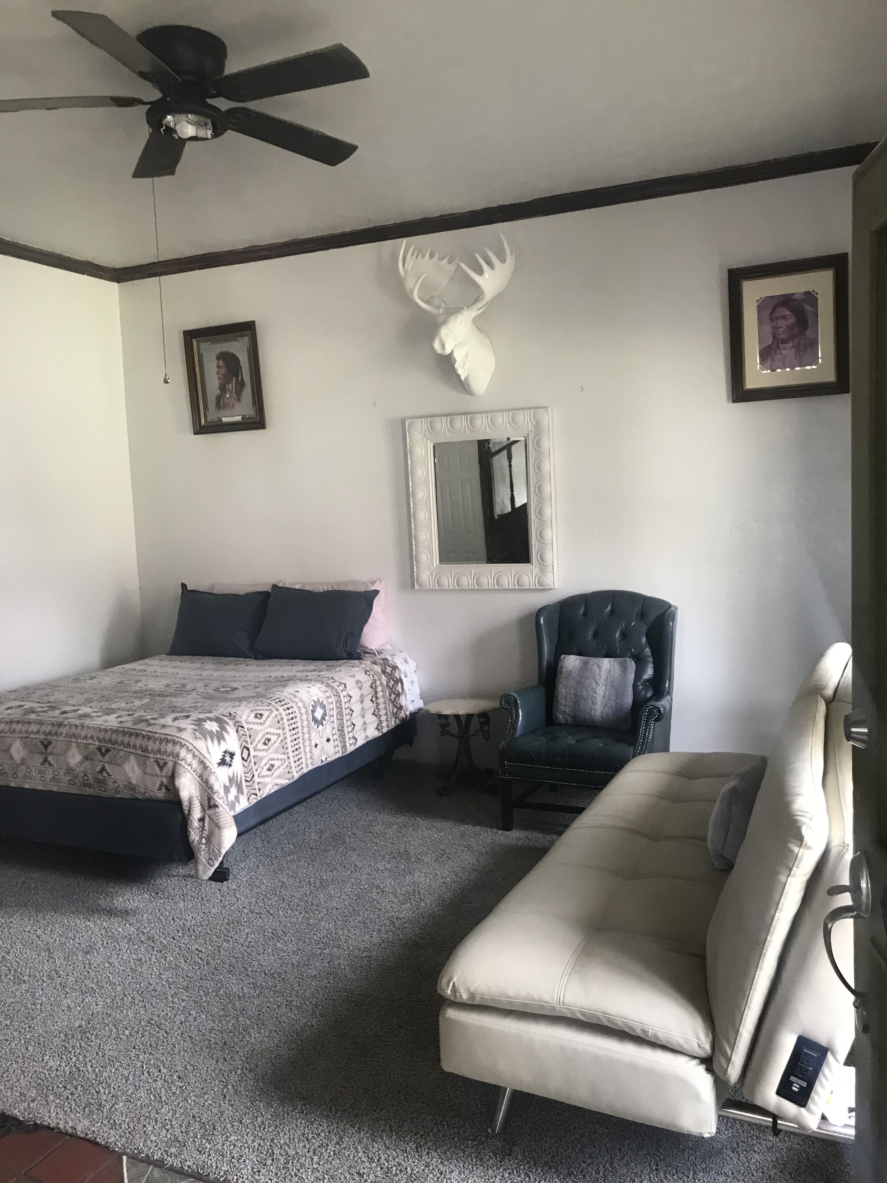 A bedroom with a bed, a chair, a sofa, a ceiling fan, a mirror, and a deer head decoration.