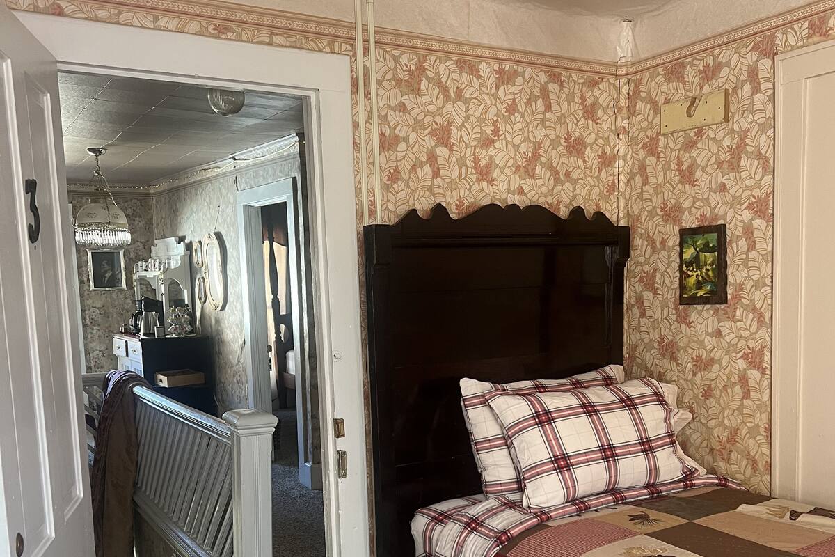 A bedroom with a bed, a wooden nightstand, a desk, and a chair. There is a door leading to another room.