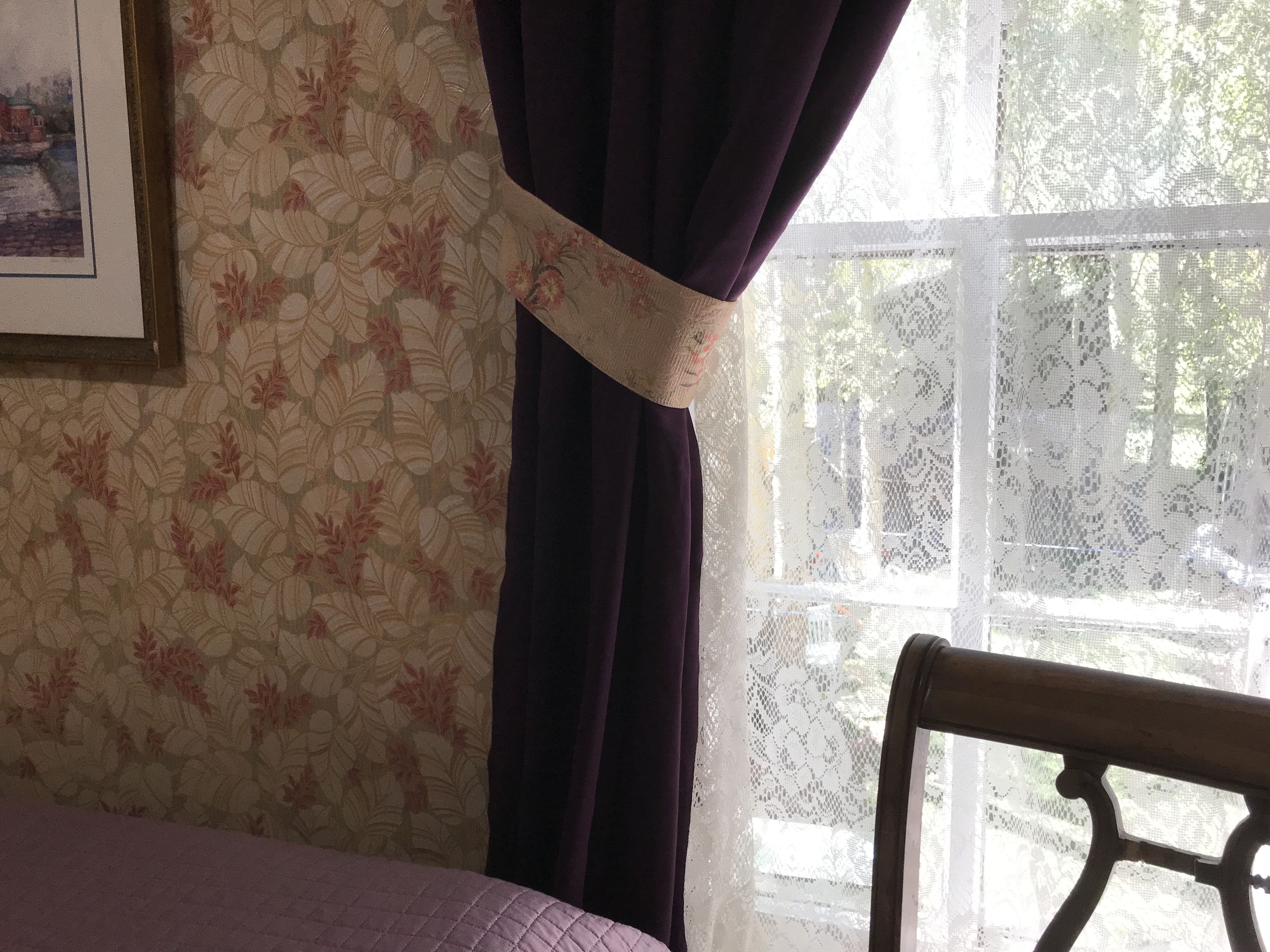 A room with floral wallpaper, a bed with a purple bedspread, a window with lace curtains, and a chair.