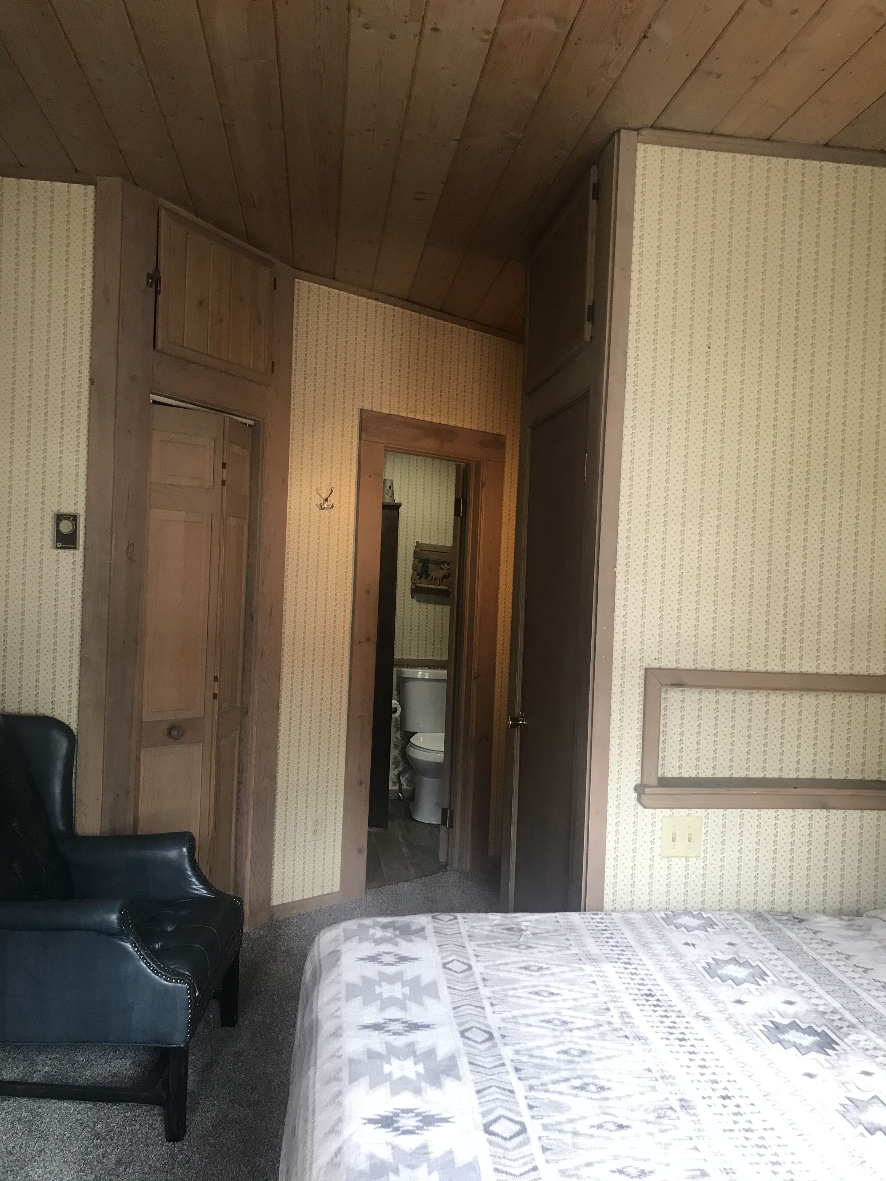 A bedroom with a bed, a wooden door, a black chair, and a bathroom visible through an open door.