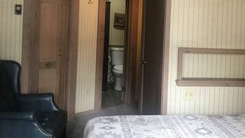 A bedroom with a bed, a wooden door, a black chair, and a bathroom visible through an open door.