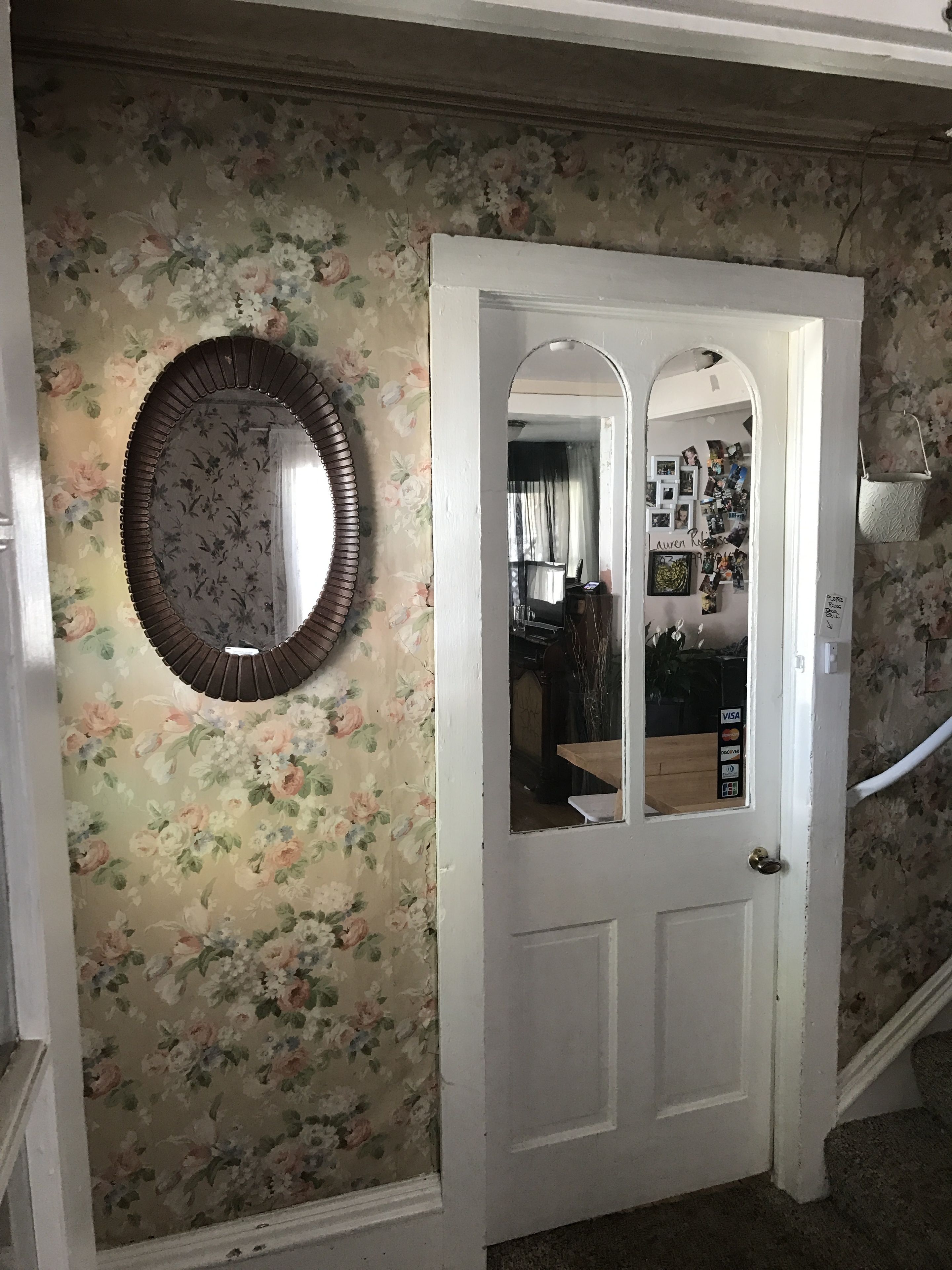 Un couloir orné d’un papier peint à motifs floraux, un miroir rond et une porte blanche avec un panneau de verre.