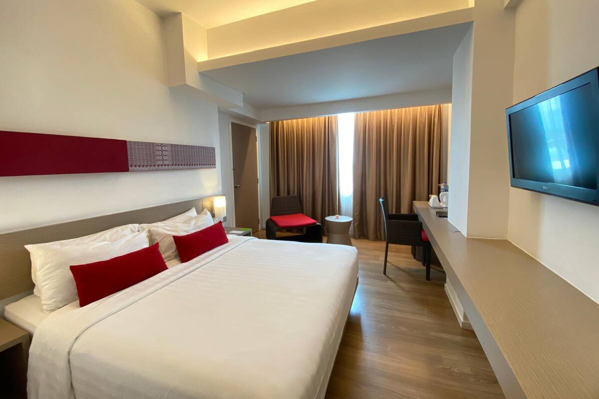 A hotel room with a large bed, a desk, a chair, a TV, and a view of the outside through a window.