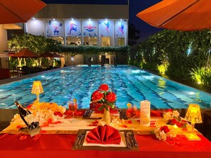 A dining table set with red napkins, cutlery, and a bottle of champagne, overlooking a swimming pool with a view of a building with illuminated artwork. - Grand Zuri Dumai (Dumai)