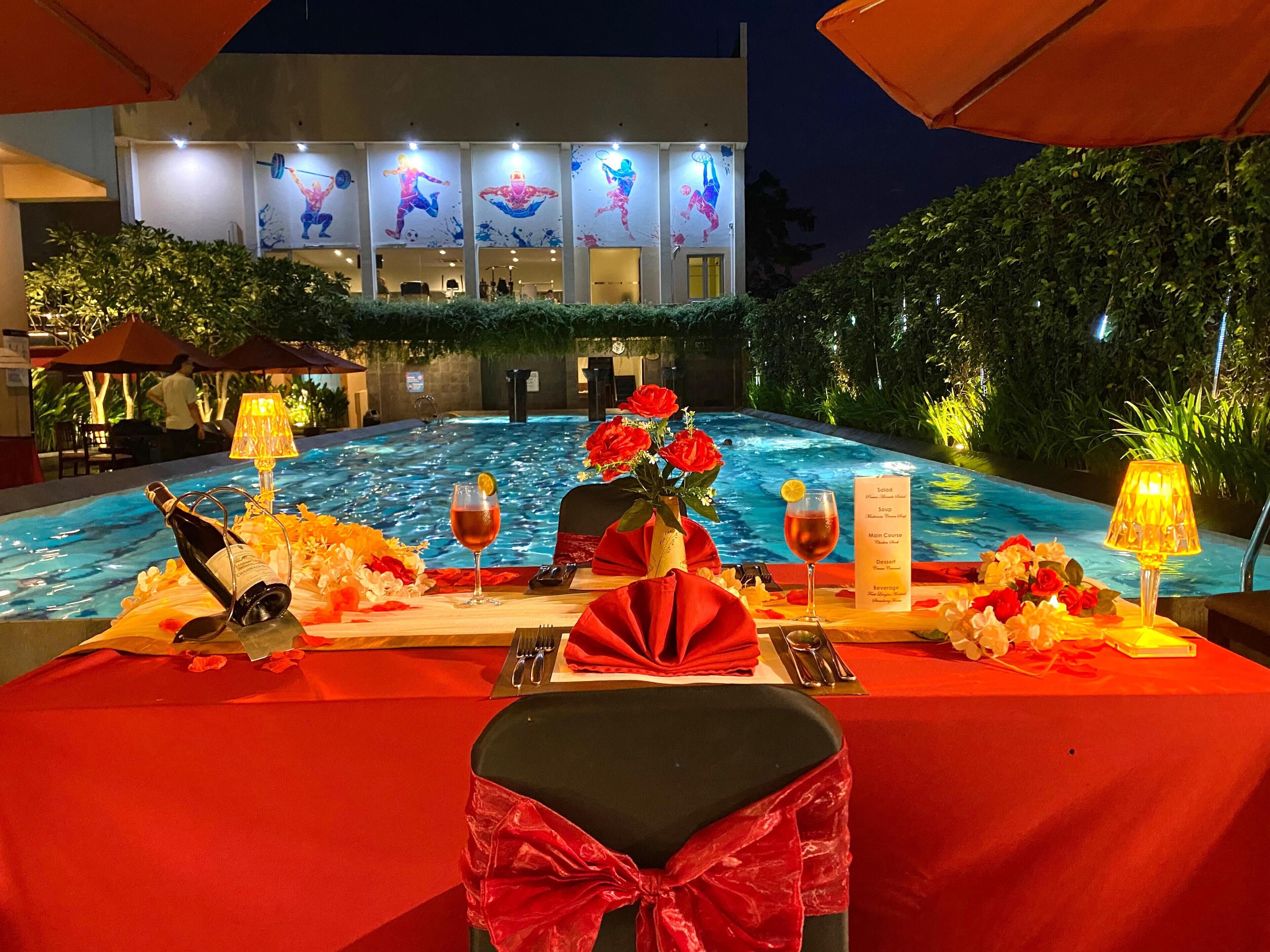 A dining table set for two with a pool view, red tablecloth, and floral decorations.
