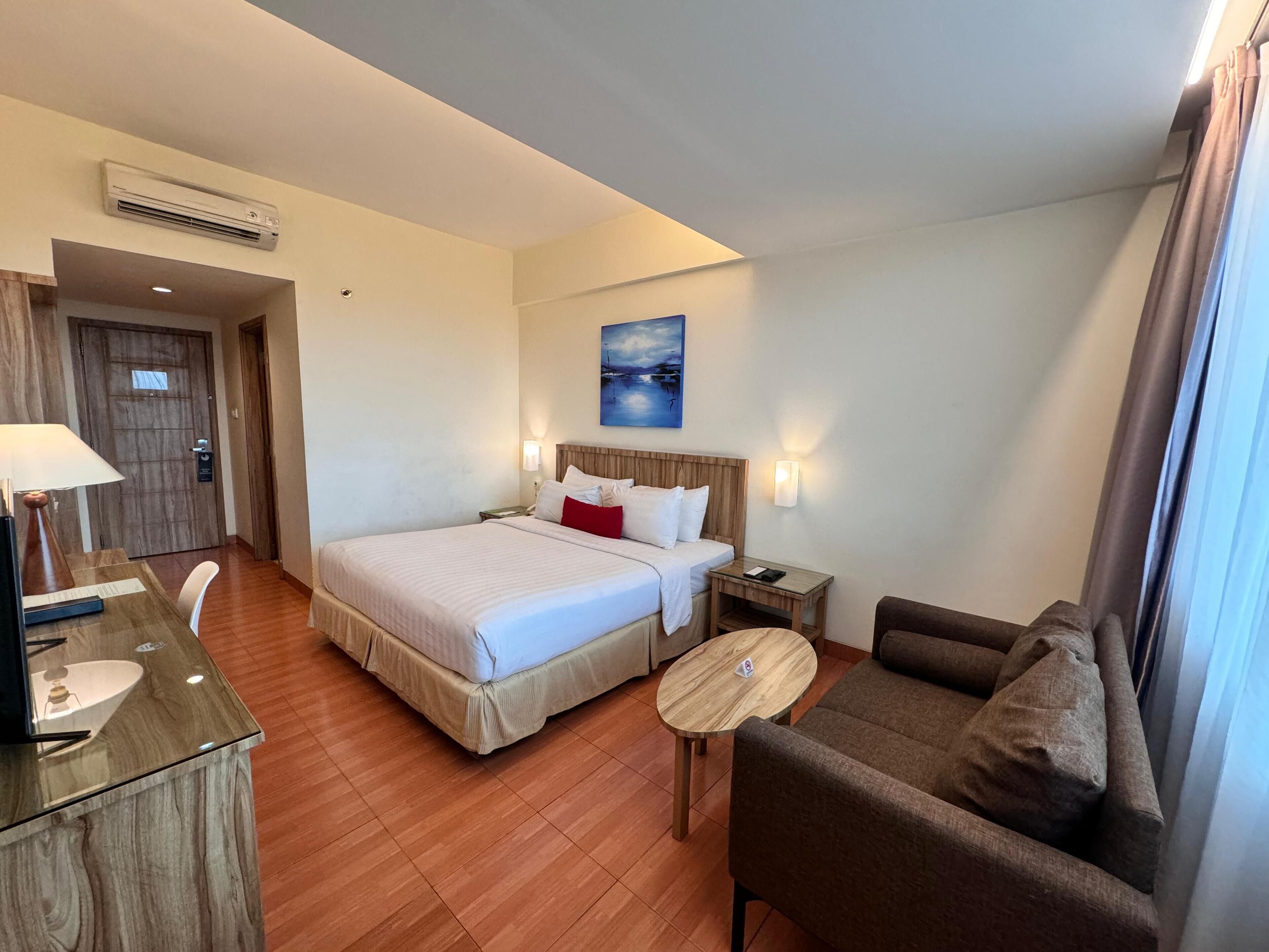 A hotel room with a bed, a desk, a chair, a sofa, and a painting on the wall.
