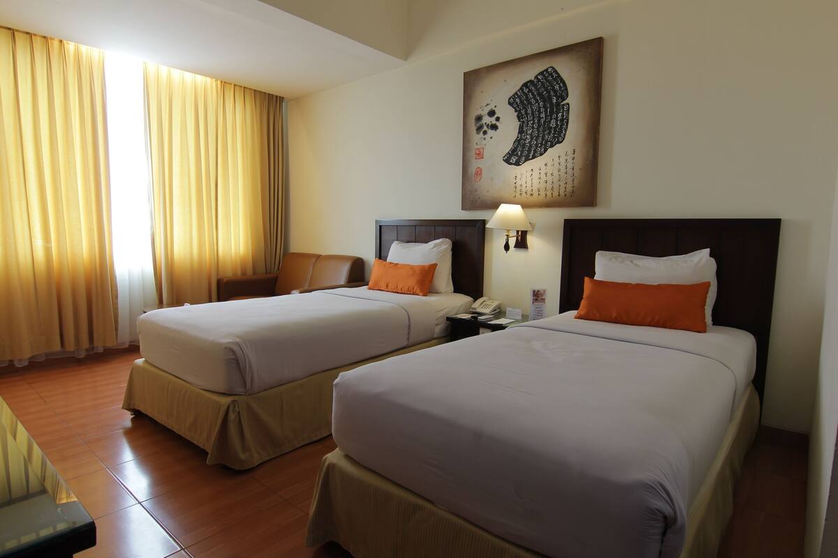 A hotel room with two beds, a desk, a chair, a lamp, and a wall-mounted artwork.