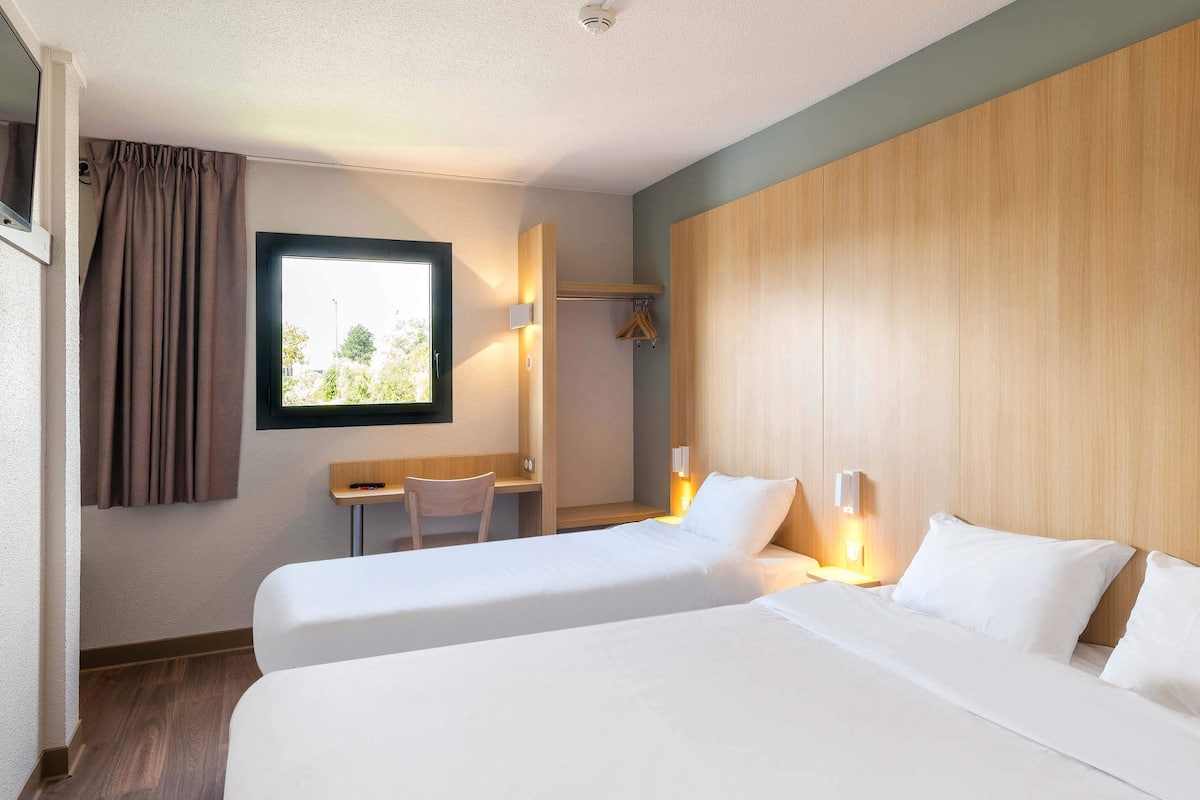 A hotel room with two beds, a desk, a chair, and a window with curtains.