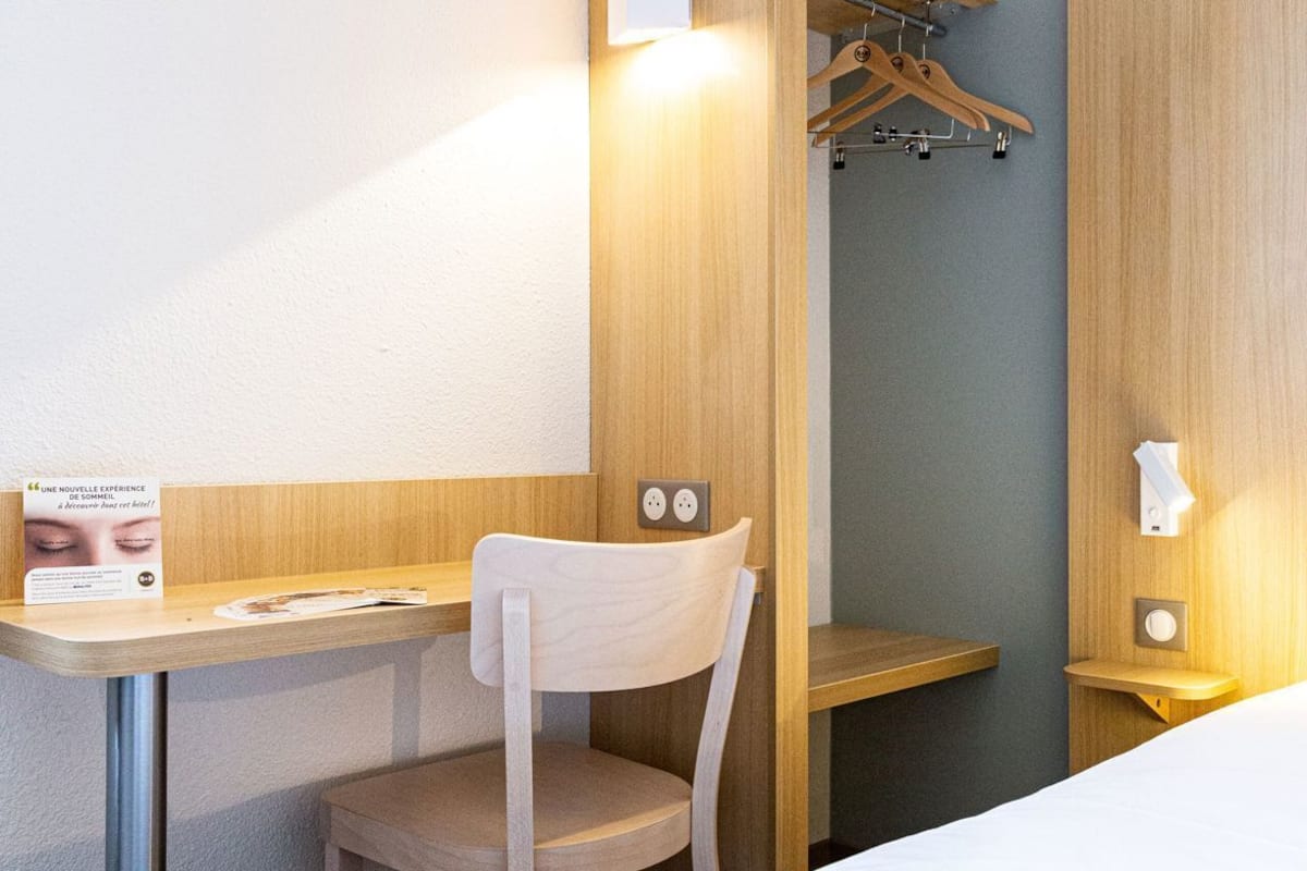 A hotel room with a wooden desk, a chair, a hanging coat rack, and a bed.