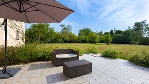 Un espacio al aire libre con patio, un sofá y reposapiés de mimbre, un paraguas grande y vistas a un campo cubierto de césped y árboles.