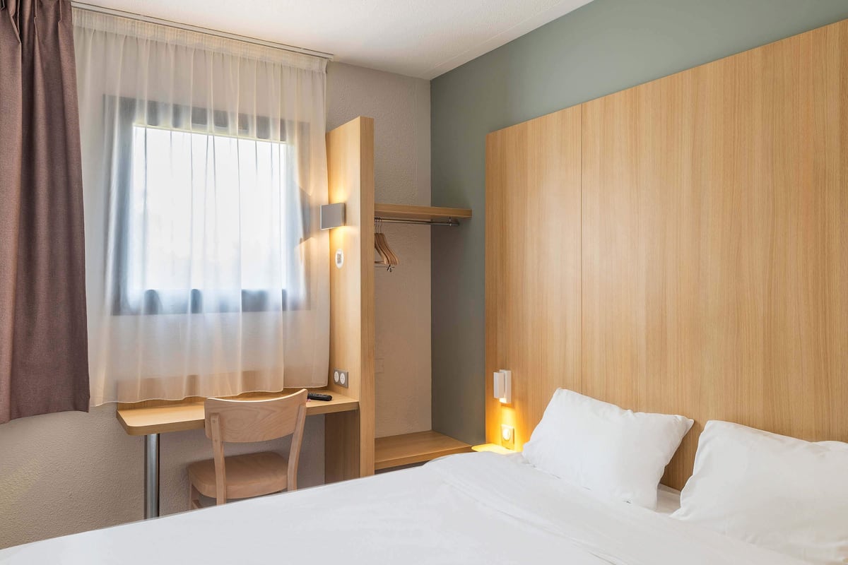 A hotel room with a bed, a desk, a chair, a shower area, and a window with curtains.
