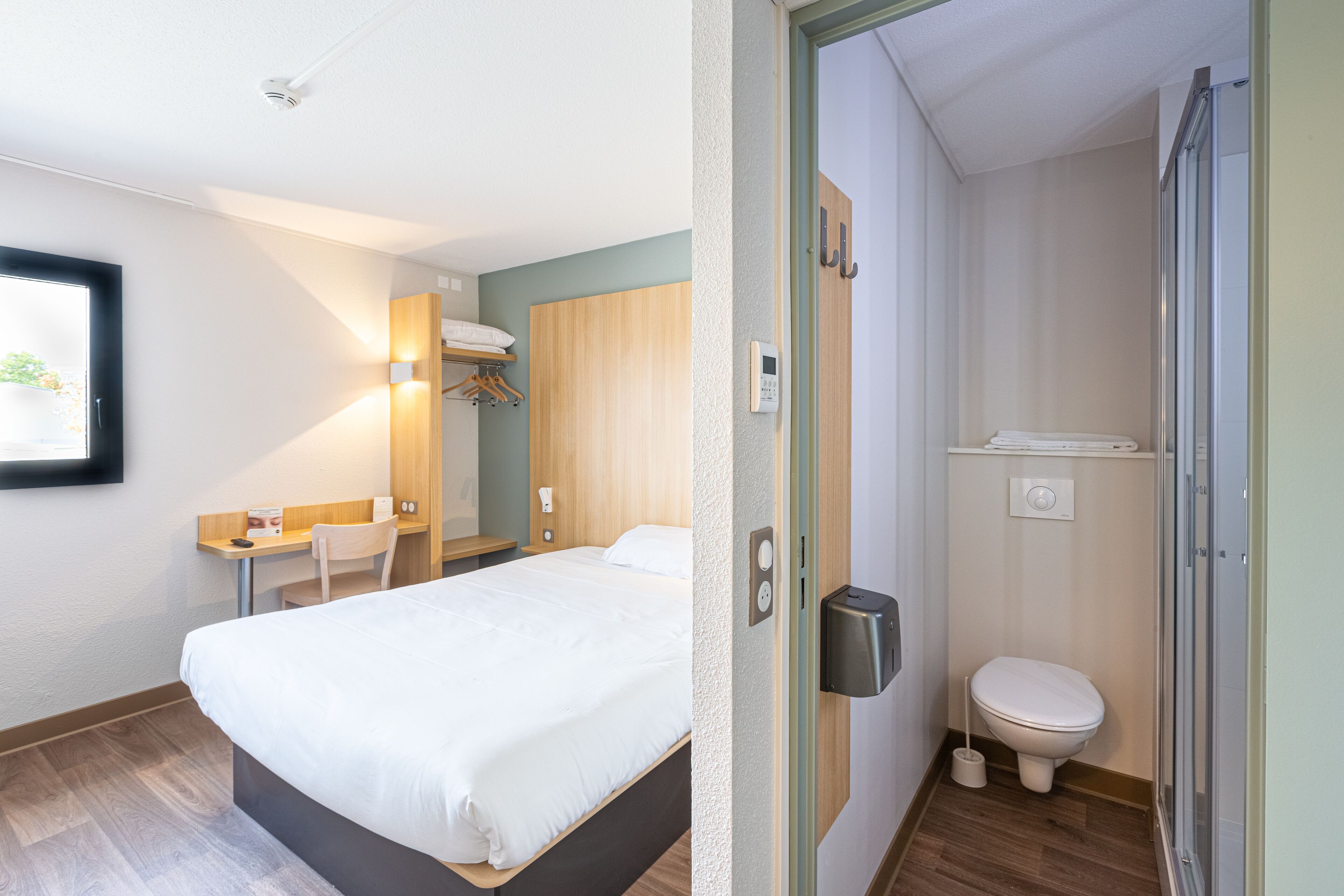 A hotel room with a bed, a desk, a chair, a wardrobe, and a bathroom with a toilet and shower.