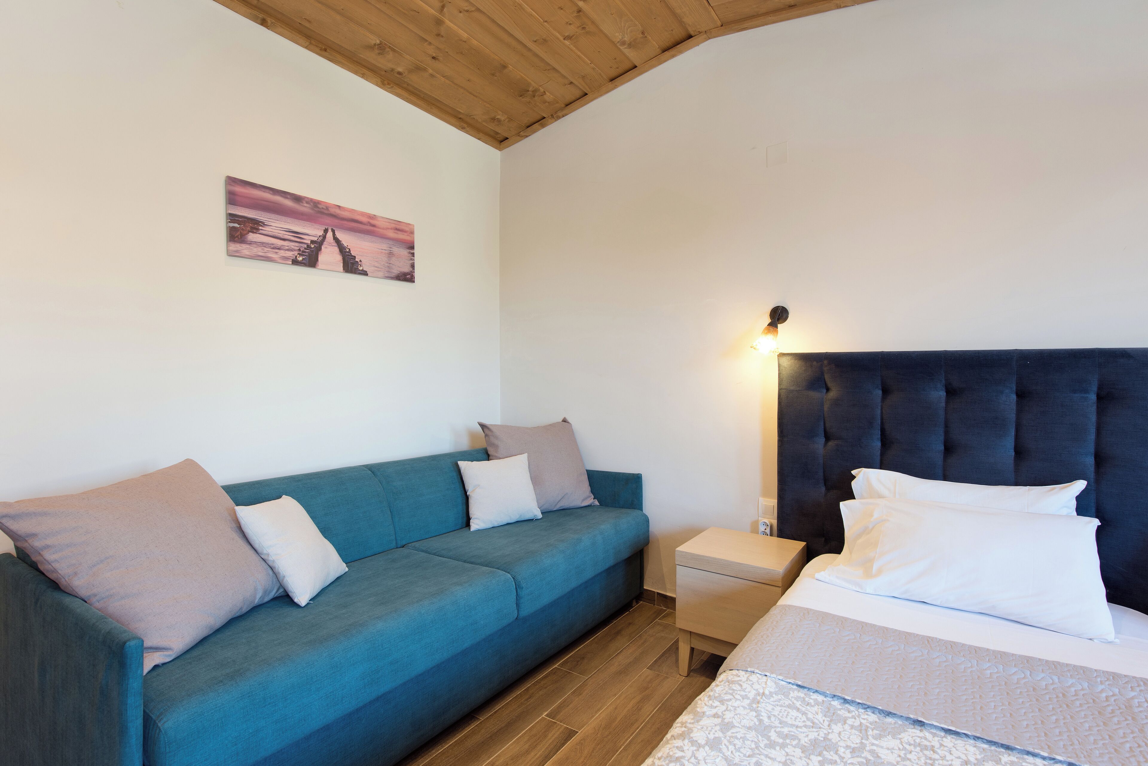 A bedroom with a blue sofa, a bed with a dark headboard, a nightstand, and a wall-mounted picture.