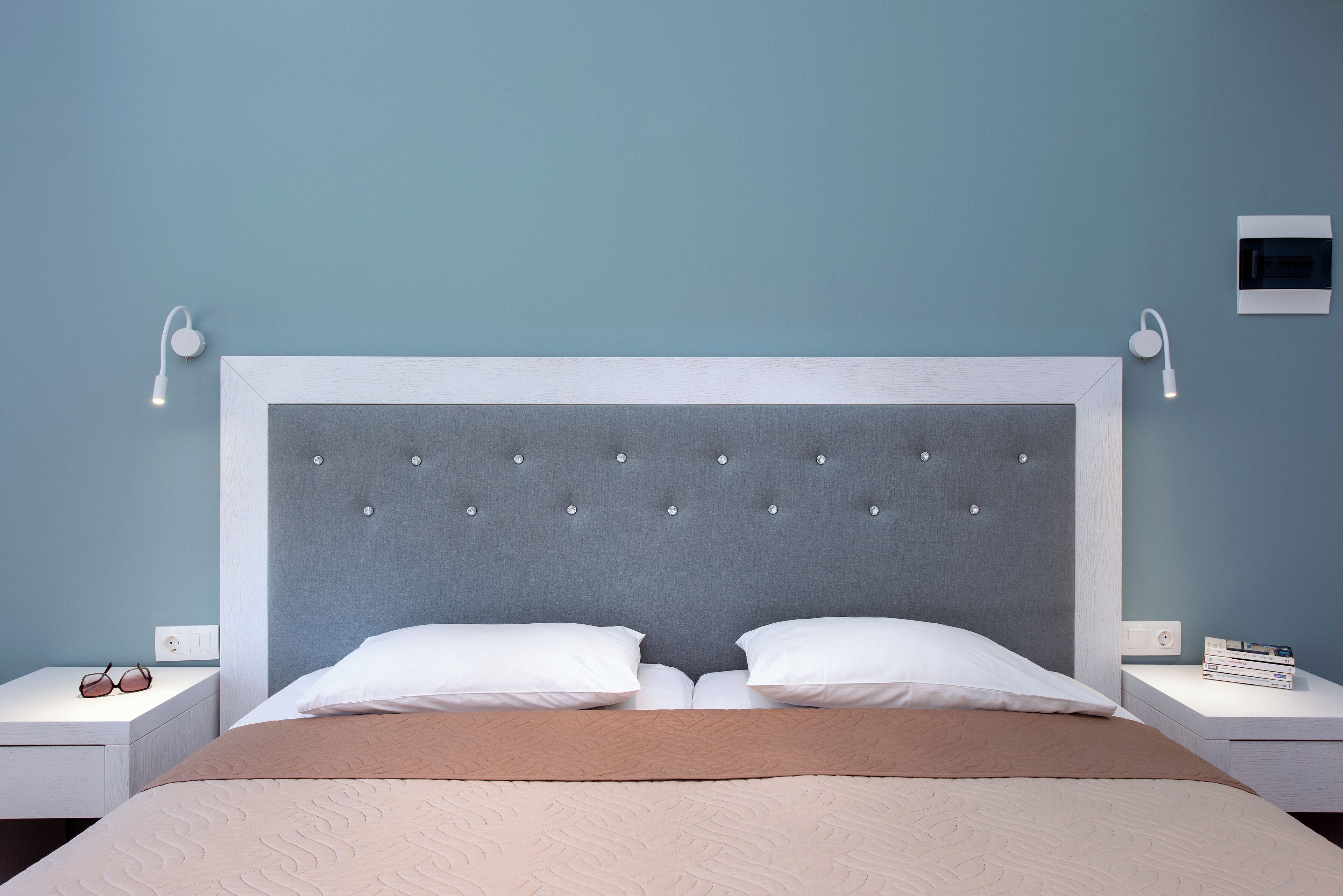 Un lit bien fait, doté d’une tête de lit capitonnée, de deux oreillers blancs et d’une couverture beige. On y trouve deux tables de chevet avec des lampes et une petite étagère avec des livres.