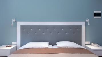 Un lit bien fait, doté d’une tête de lit capitonnée, de deux oreillers blancs et d’une couverture beige. On y trouve deux tables de chevet avec des lampes et une petite étagère avec des livres.