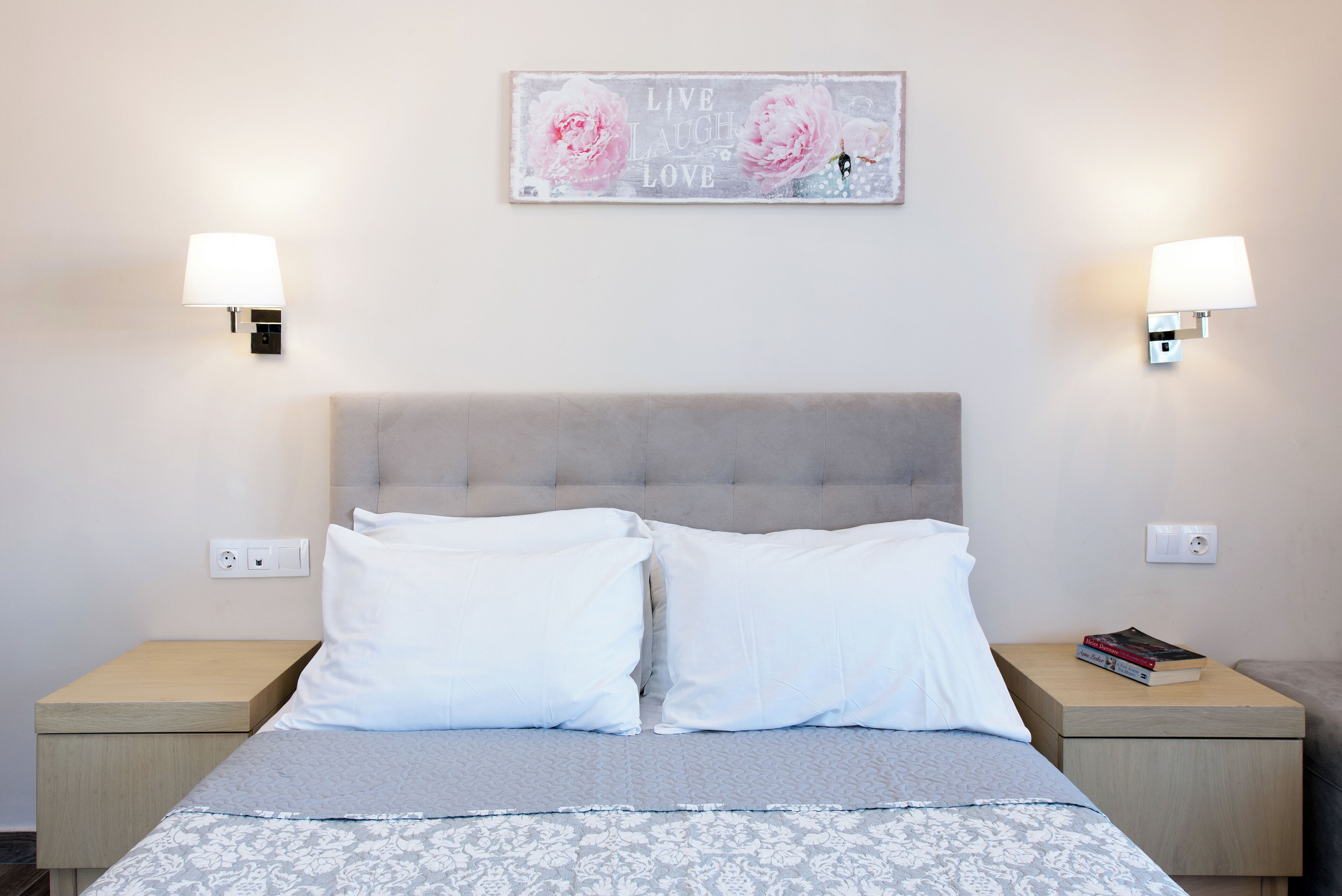 A neatly made bed with white and grey bedding, two bedside tables with lamps, a wall-mounted light switch, and a decorative sign above the bed.