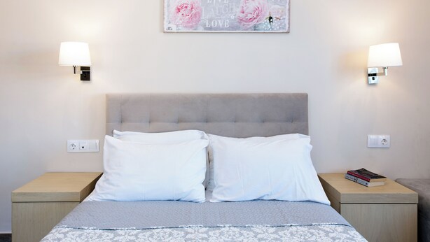 A neatly made bed with white and grey bedding, two bedside tables with lamps, a wall-mounted light switch, and a decorative sign above the bed. - Anita Hotel (Corfu)