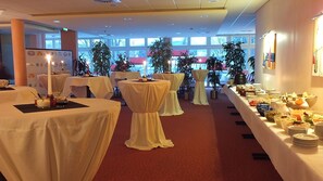 A banquet hall with round tables covered in white tablecloths, a buffet line with various food items, and potted plants. - Fletcher Parkhotel Olsberg-Winterberg (Olsberg)