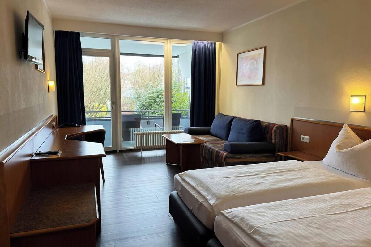 A hotel room with a bed, a desk, a chair, a sofa, and a large window with curtains.
