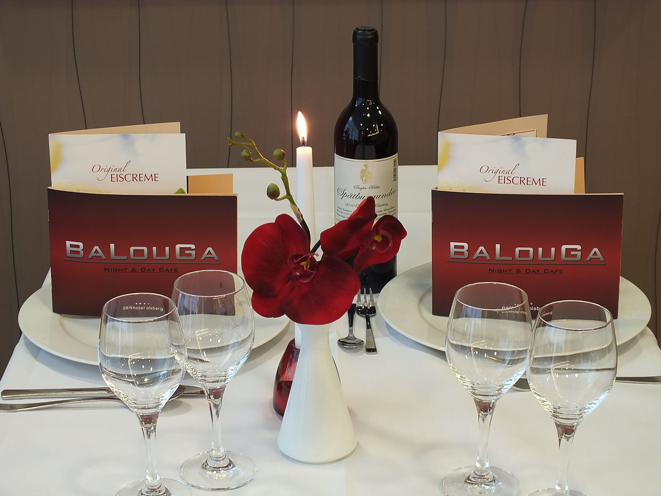 Ένα τραπέζι στρωμένο με κουτιά από το Balouga Night & Day Cafe, ένα μπουκάλι κρασί, ένα αναμμένο κερί και αρκετά ποτήρια κρασιού.