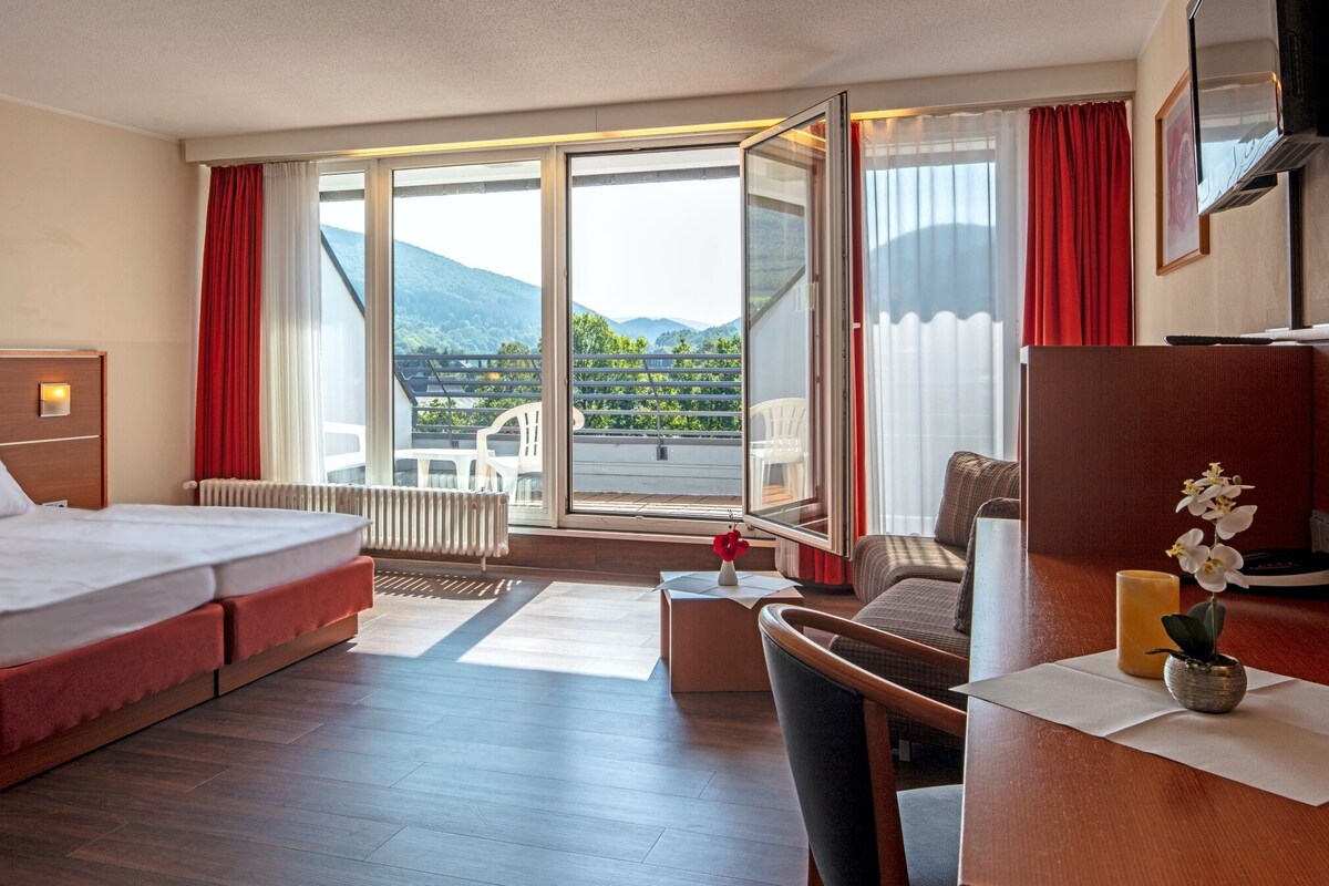 A hotel room with a bed, a desk, a chair, a small table, a vase with flowers, a radiator, and a large window with a view of mountains.