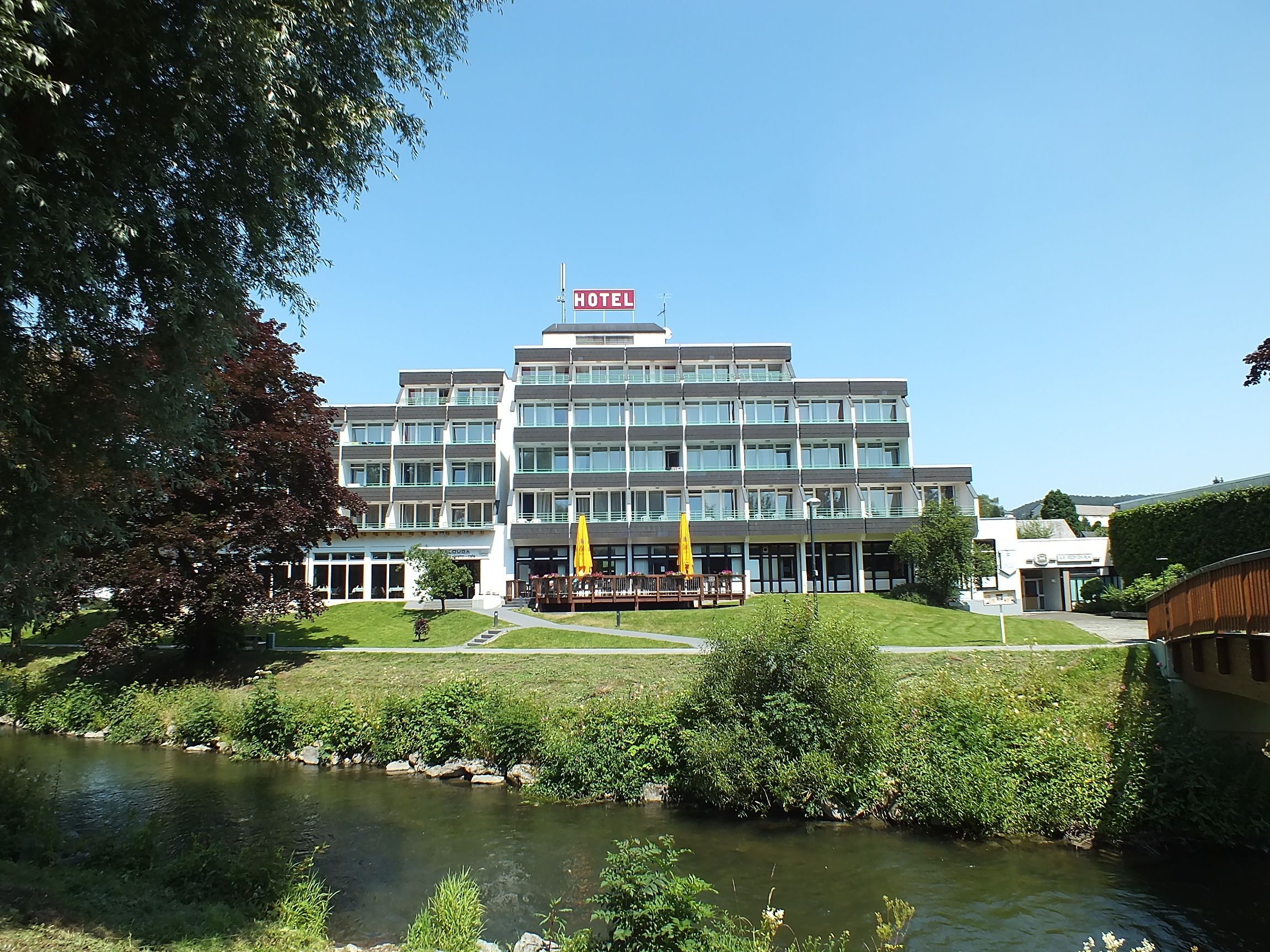 Un hôtel à plusieurs étages, surmonté d’une enseigne lumineuse indiquant « HÔTEL », avec une cour verdoyante et un petit ruisseau au premier plan.