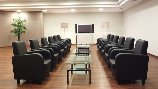 A conference room with rows of black chairs, a glass table, a large flat-screen TV, and two floor lamps. - Fletcher Parkhotel Olsberg-Winterberg (Olsberg)