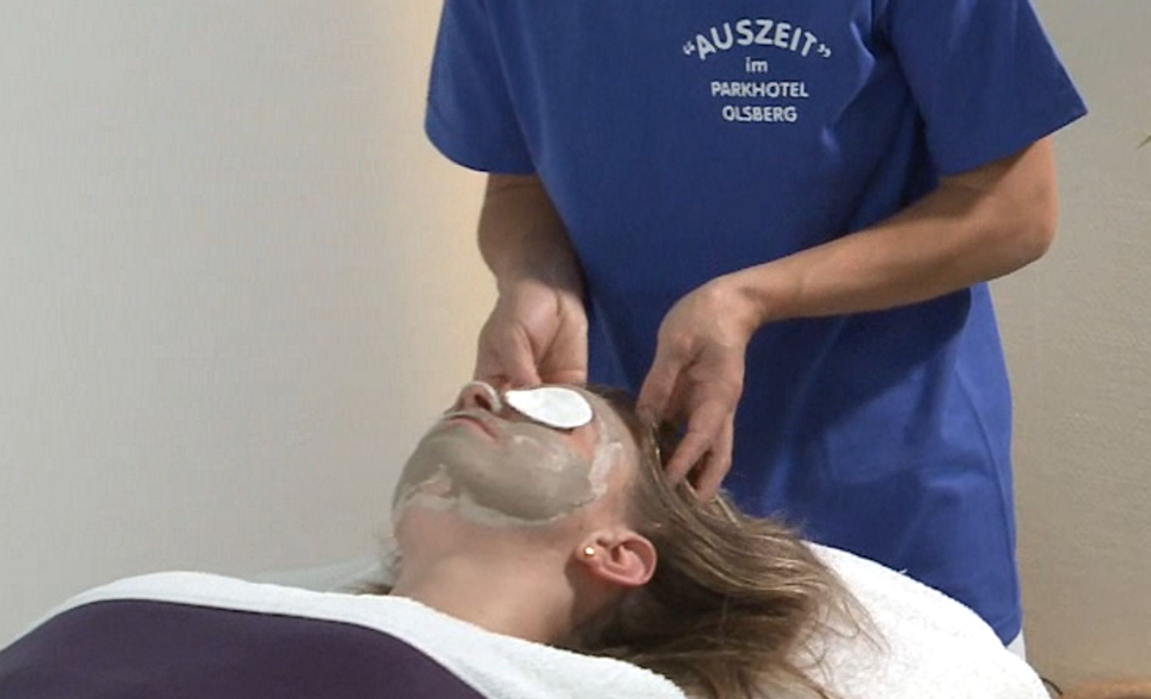 A person receiving a facial treatment with a white mask, wearing a blue shirt with text, in a room with a plain wall.