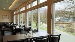 A dining area with tables and chairs, large windows, and a view of a bridge and greenery. - Fletcher Parkhotel Olsberg-Winterberg (Olsberg)