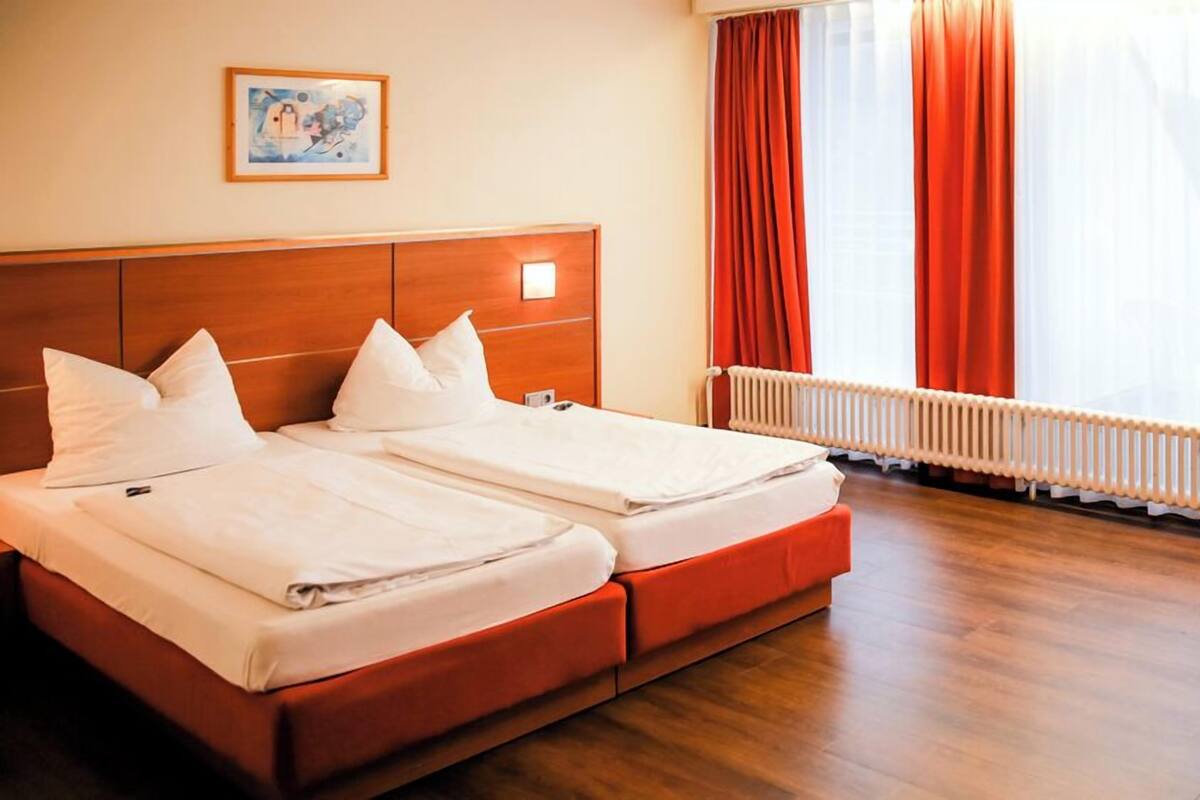 A hotel room with two beds, a wooden headboard, a radiator, and a window with curtains.