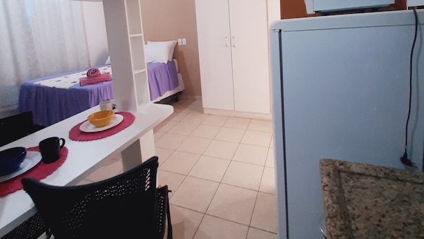 Uma cozinha compacta com geladeira branca, uma mesa de jantar com cadeiras e uma cama com manta roxa.