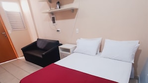 Quarto de hotel com uma cama, mesa de cabeceira, cadeira e uma estante com alguns objetos.