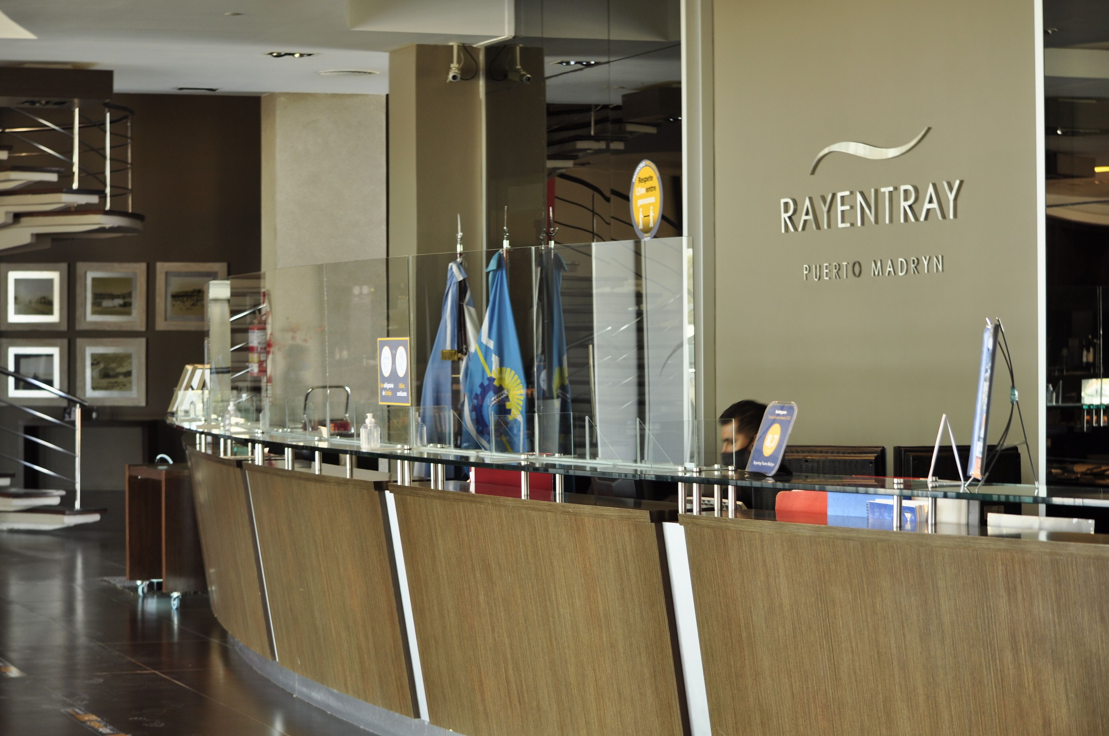A reception desk with a glass partition, a sign reading "RAYENTRAY PUERTO MADRYN", and a staircase in the background.