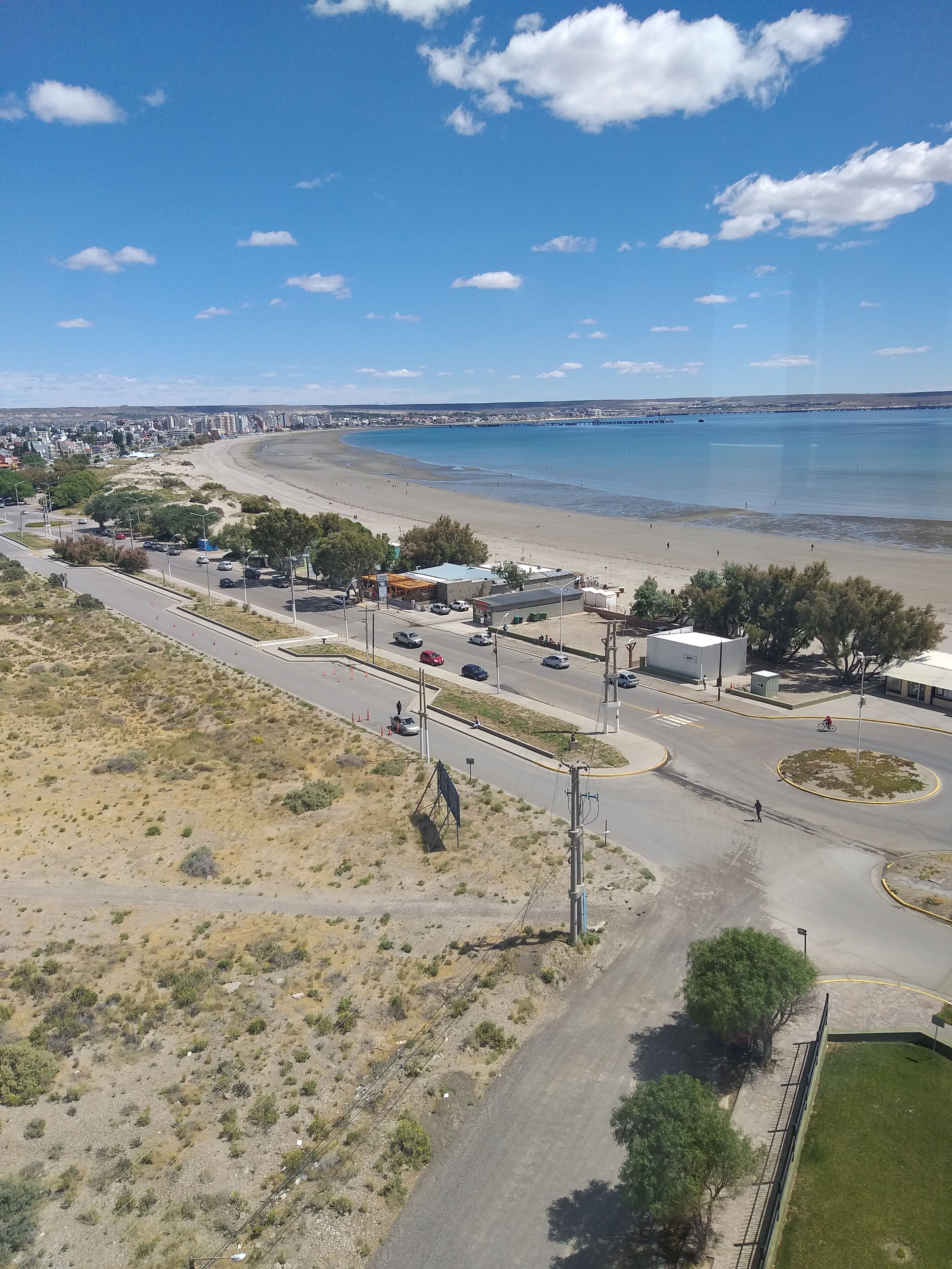 An aerial view of a coastal area with a beach, a road with vehicles, and buildings in the background.