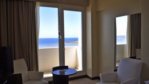 Hotelkamer met een grote schuifglazen deur die uitkomt op een balkon met uitzicht op zee. Er staan twee witte fauteuils en een klein rond tafeltje met een tijdschrift erop.