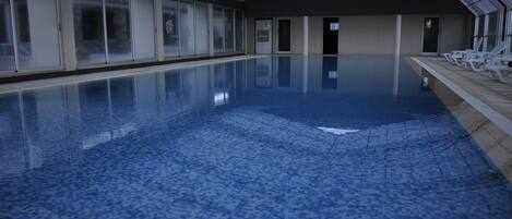 Una piscina interior con paredes de vidrio transparente, suelo de baldosas y sillones blancos a un lado.