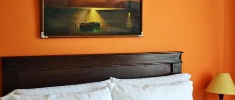 Hotelkamer met een bed, witte kussens, een schilderij van een zonsondergang boven water en een lamp op een nachtkastje.
