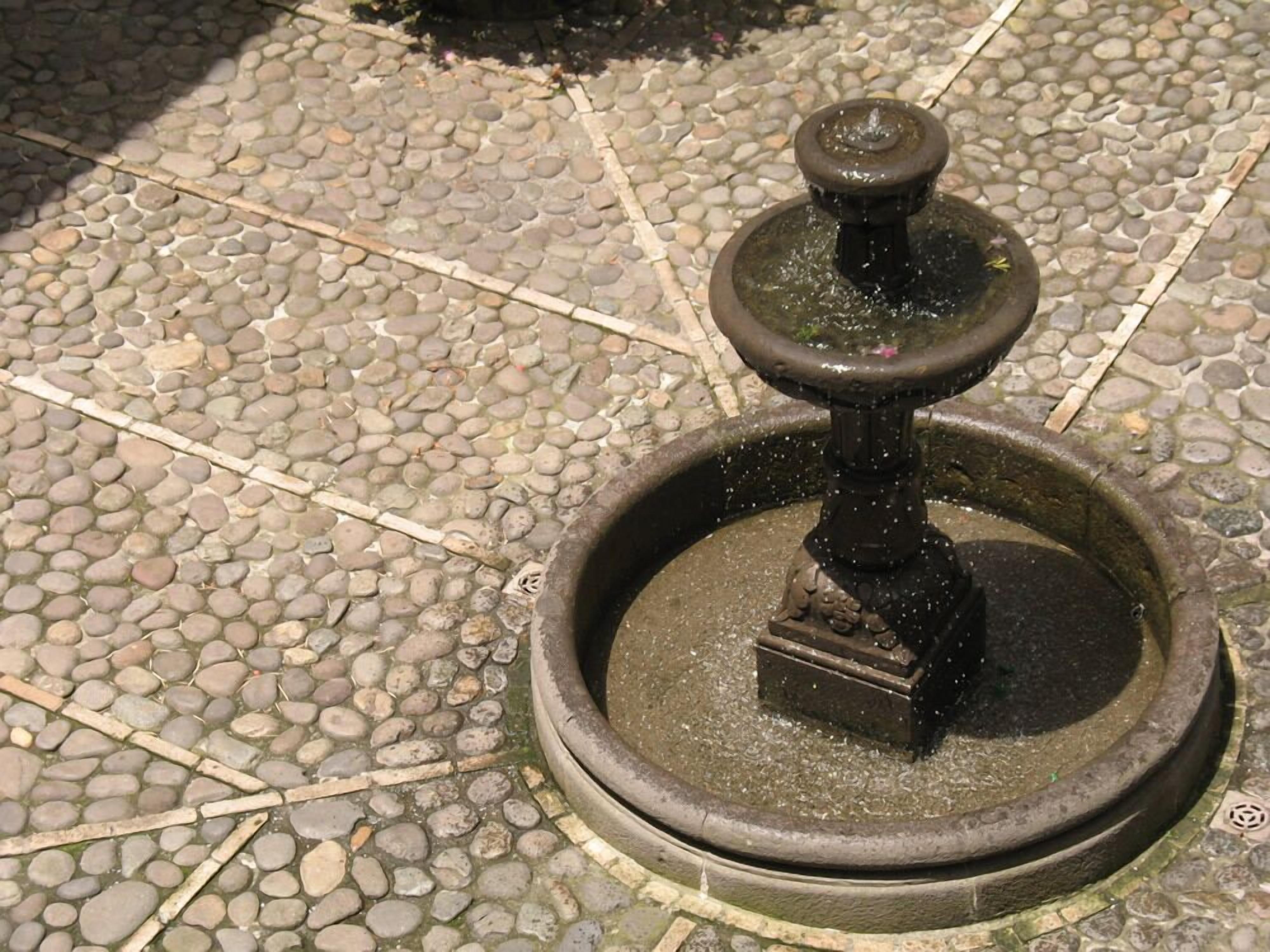 A stone fountain with a central spout and a smaller spout on top, surrounded by a circular stone base.