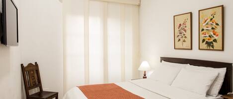 A hotel room with a bed, a chair, a TV, and two paintings on the wall.