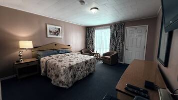 A hotel room with a bed, a desk, a chair, a lamp, a television, and a door.