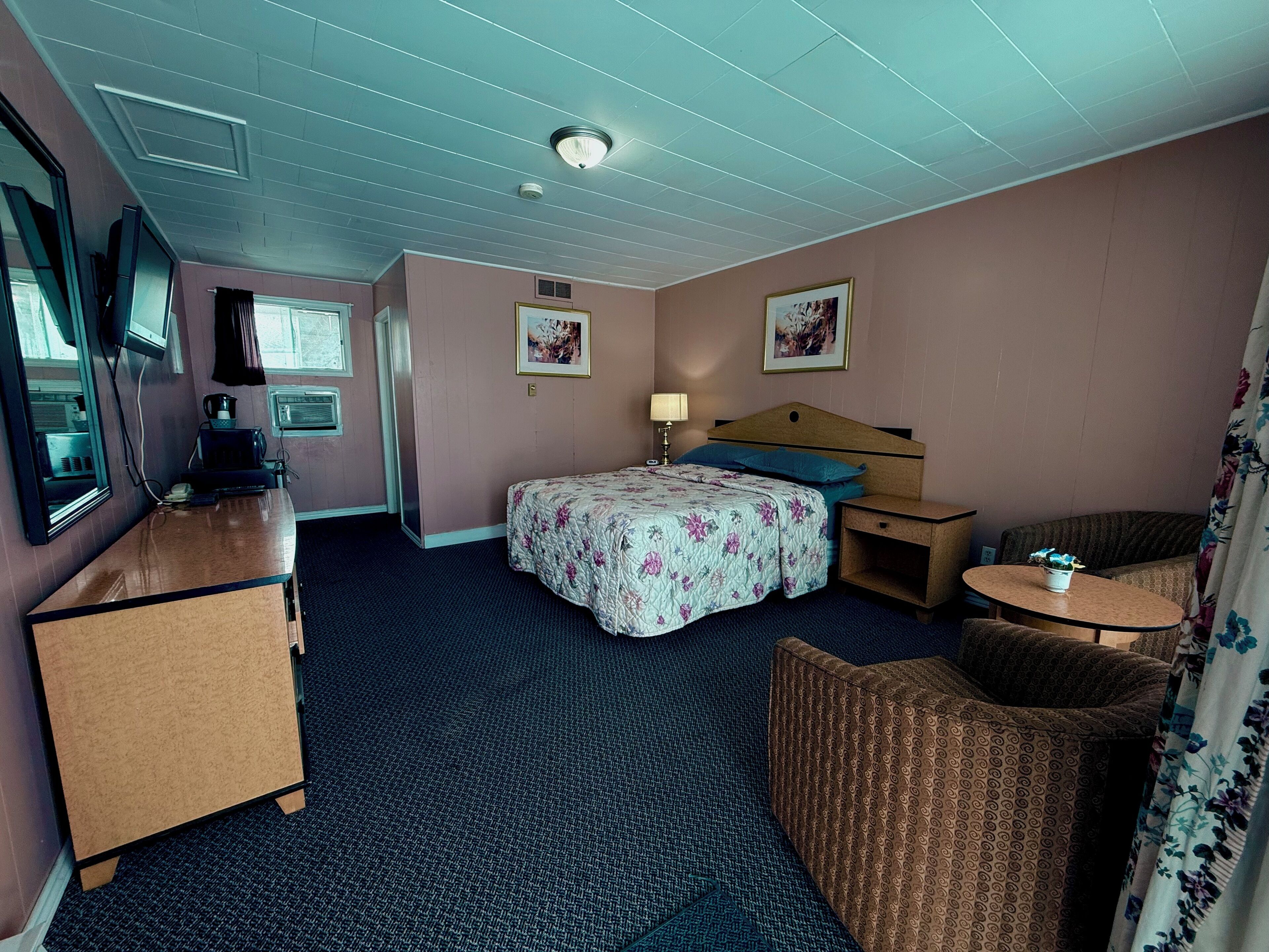 A hotel room with a bed, a desk, a chair, a small table, a TV, and a window.