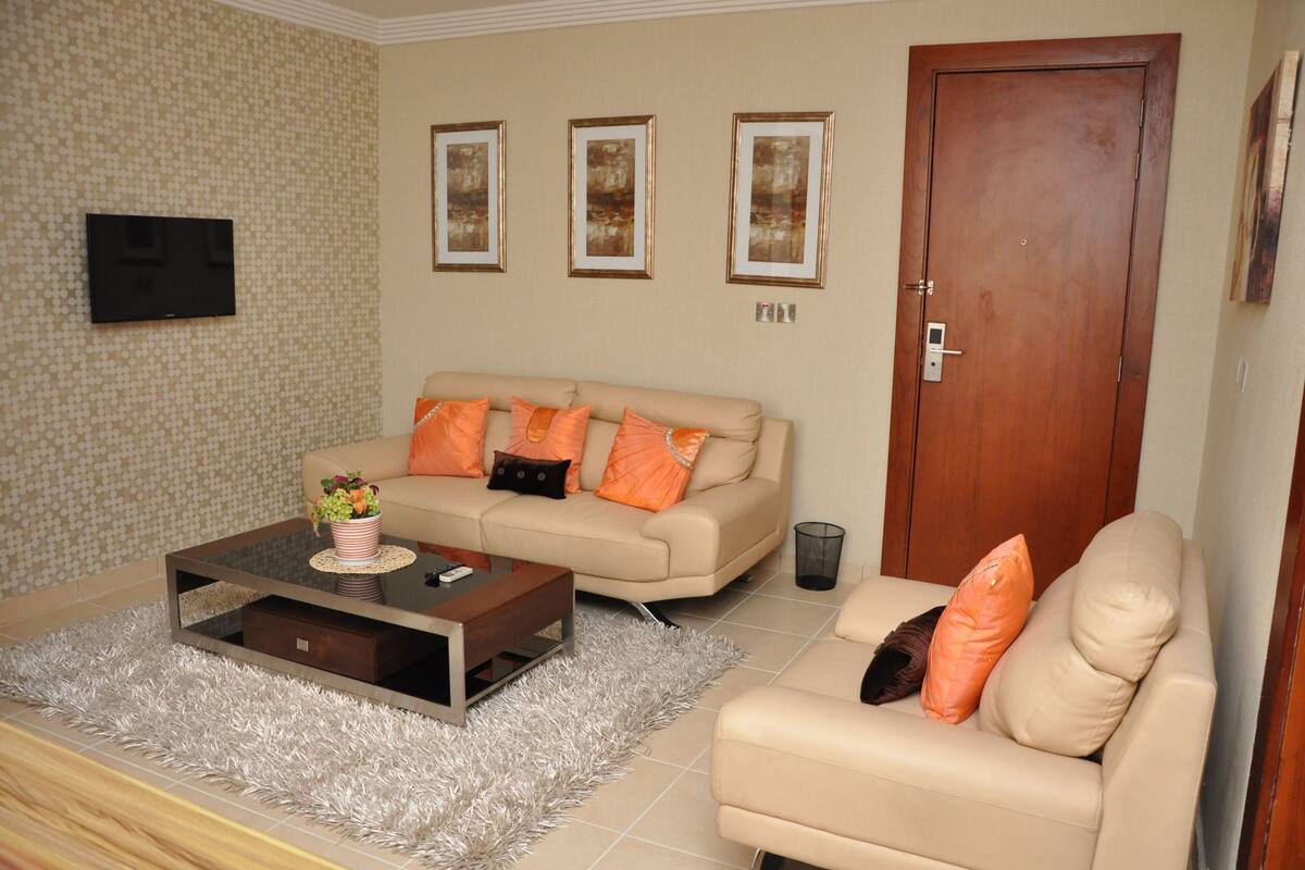 Ein Wohnzimmer mit einem beigefarbenen Sofa, orangefarbenen Kissen, einem Glastisch und einem an der Wand montierten Fernseher.
