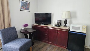 Una habitación de hotel compacta con televisión, cafetera, microondas, lámpara, una mesita con un jarrón de flores, una silla y un armario.